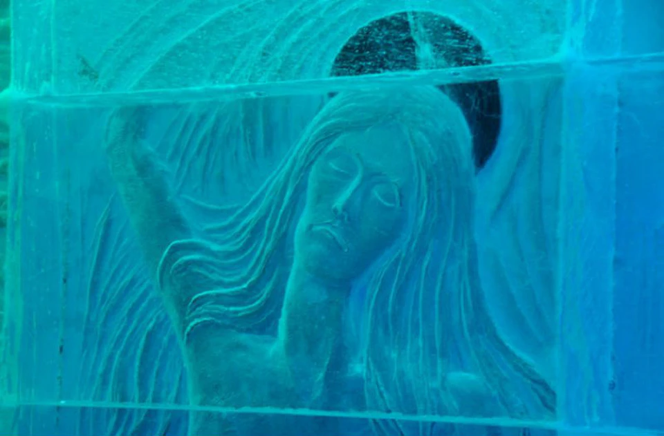 An intricate ice carving of a serene woman with long hair and a halo-like circle behind her head in a blue tint.