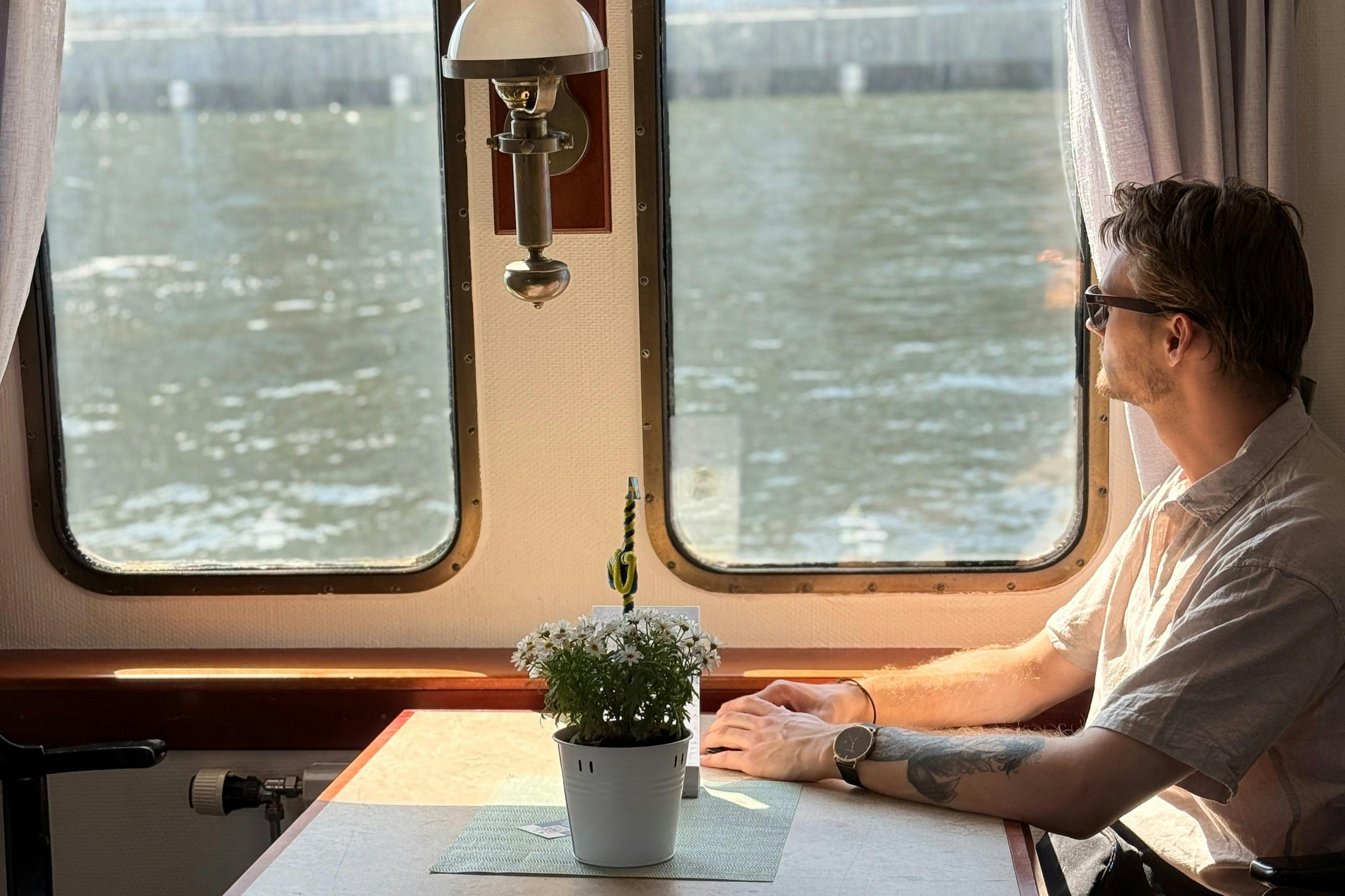 Una persona con un braccio tatuato siede a un tavolo con una pianta in vaso, vicino a una finestra che si affaccia sull'acqua.