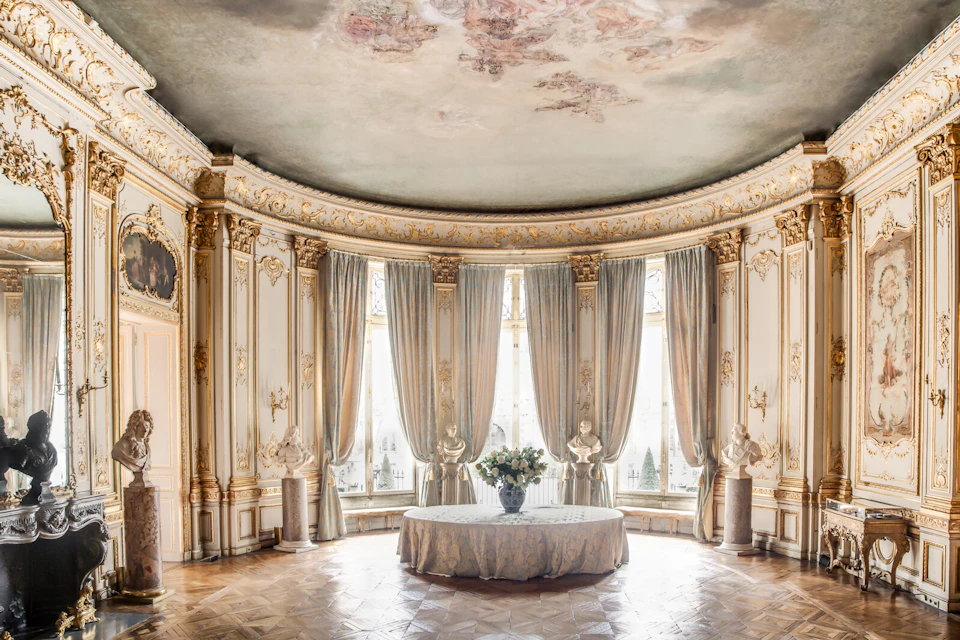 Elegant rotunda with ornate gold details, tall windows draped with curtains, statues, a round table with a floral centerpiece.