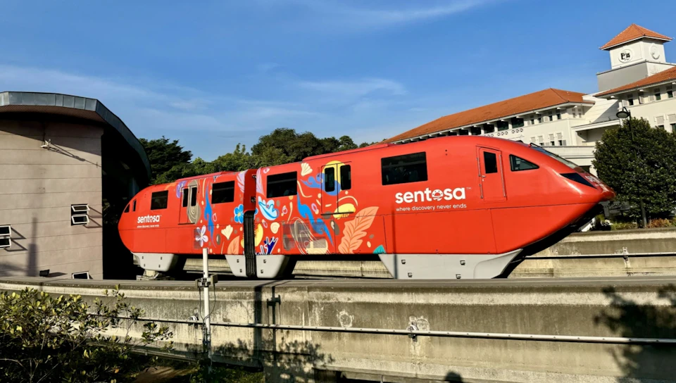 A red monorail train with colorful designs and the text 'Sentosa' on its side, moving on an elevated track under a clear sky.