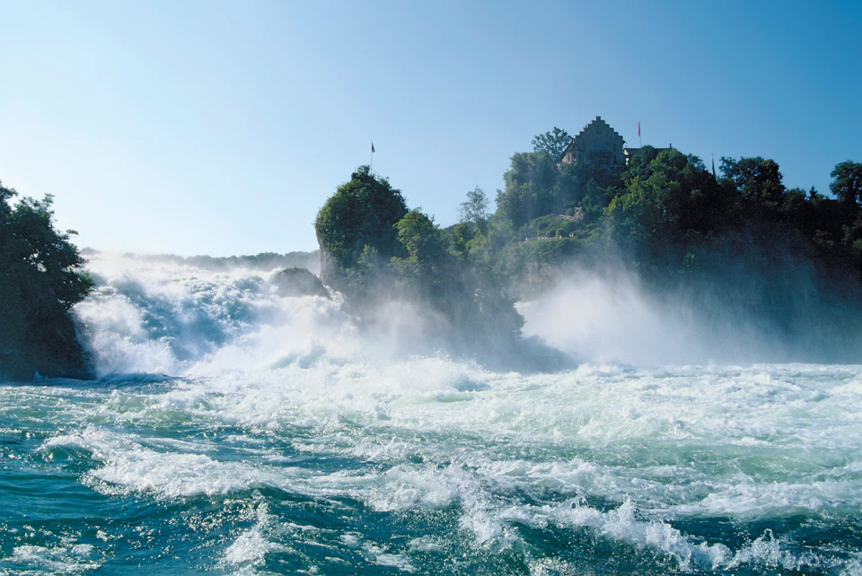 Powerful waterfall surrounded by mist and lush greenery with a large, castle-like building on a hill in the background.