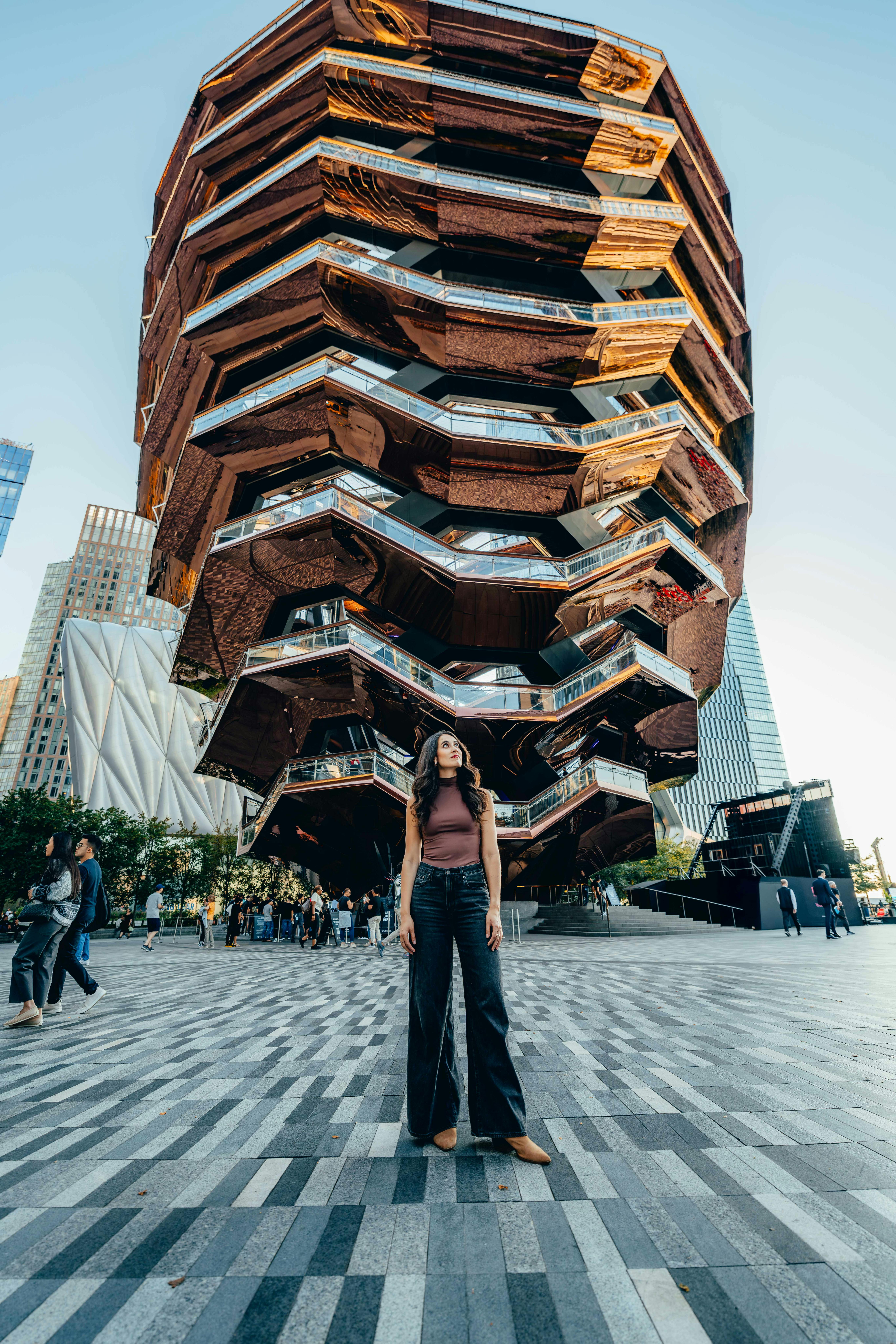 Woman standing in front of the Vessel, a honeycomb-like structure, with surrounding people and buildings in the background.
