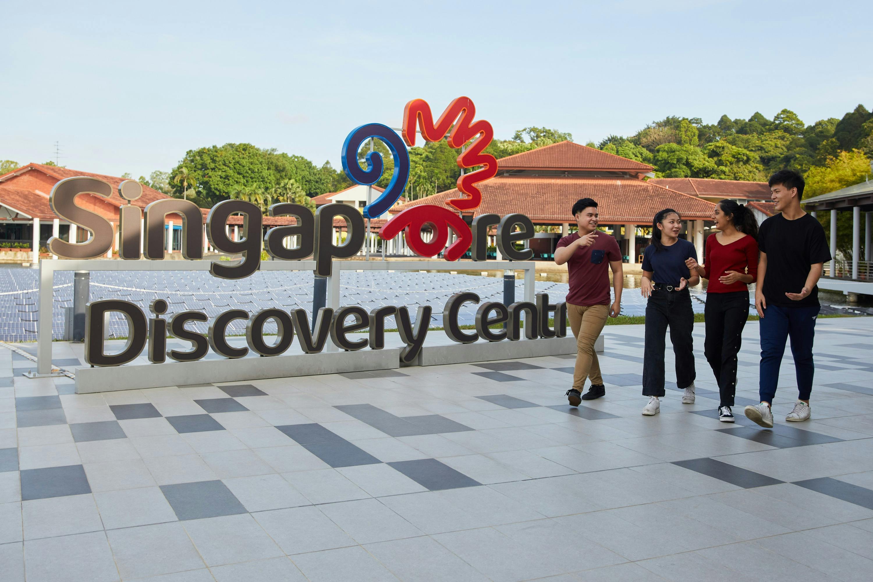 Four people walking and talking outside the Singapore Discovery Centre, with a red and blue sign in the background.