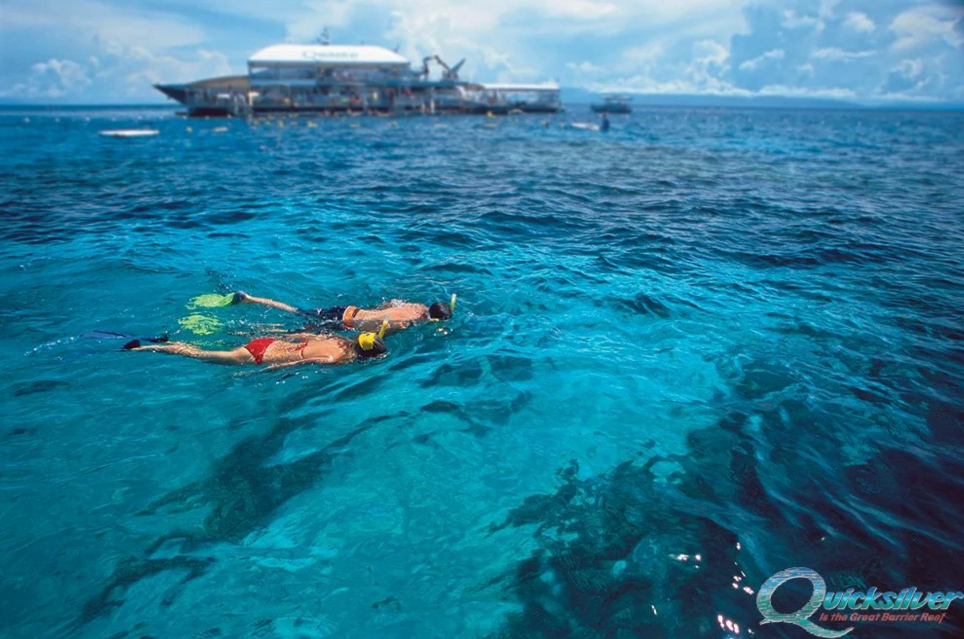 Two snorkelers in clear blue ocean water near a floating platform with equipment under a sunny sky.