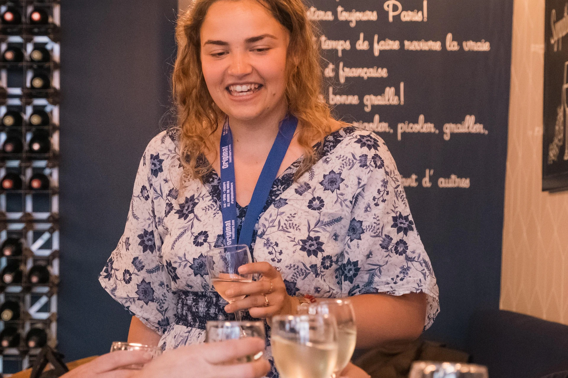 A woman smiles while holding a glass of wine, surrounded by others toasting with wine glasses in a cozy setting with wine racks.