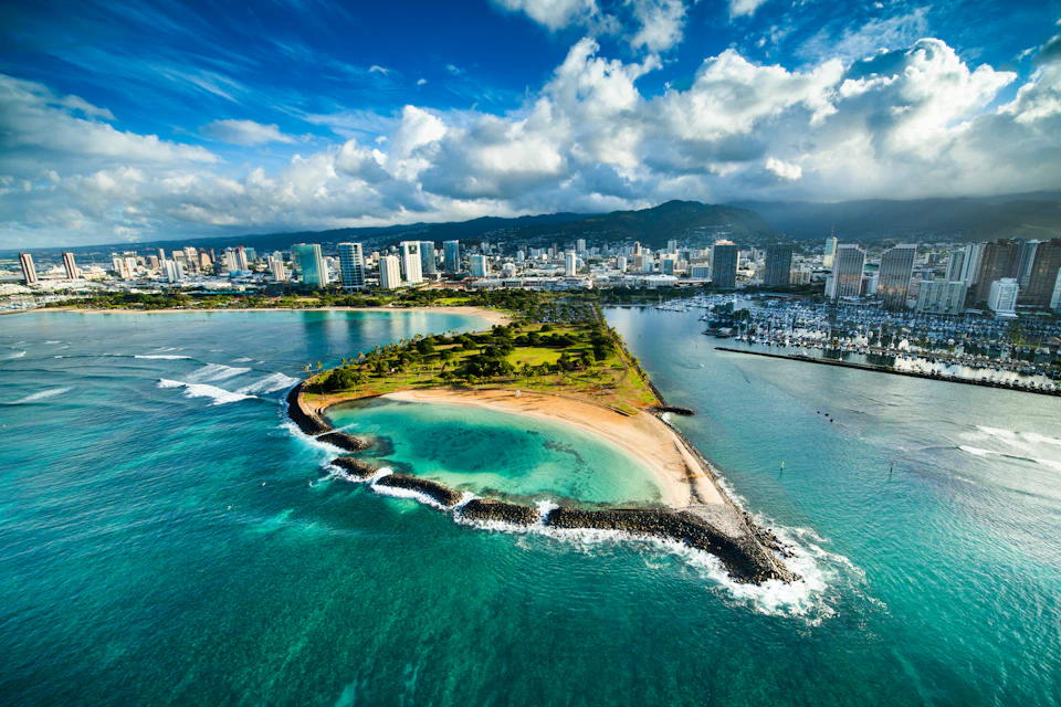 Aerial view of a city skyline and marina adjacent to a small green peninsula with a sandy beach, surrounded by turquoise water.
