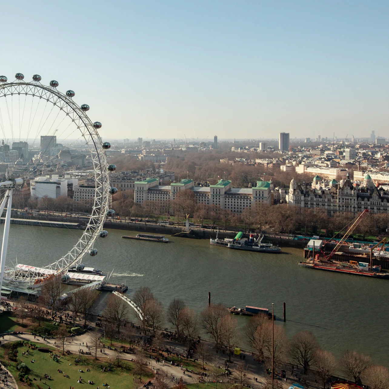 Aerial view of the London Eye beside the River Thames with surrounding buildings and a park in the foreground.