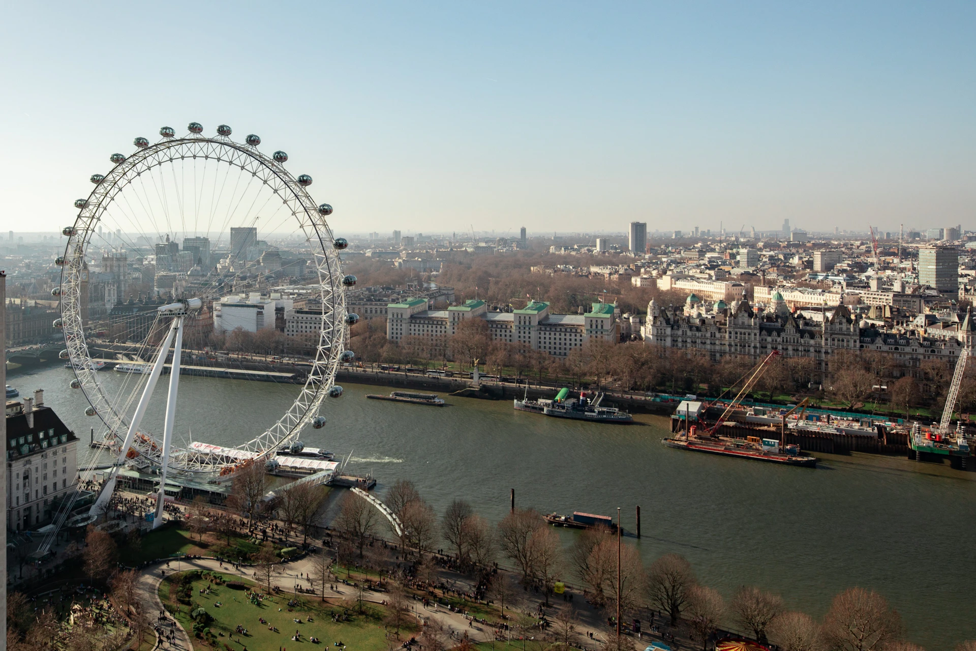 Aerial view of the London Eye beside the River Thames with surrounding buildings and a park in the foreground.