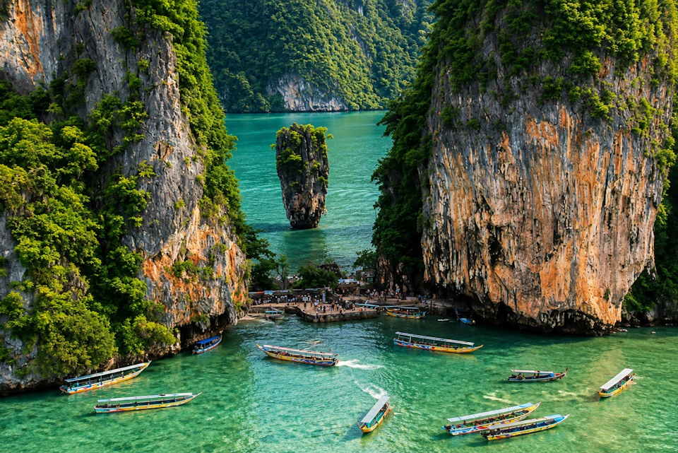 A scenic tropical bay with tall limestone cliffs, turquoise water, boats, and a vertical rock formation in the center.