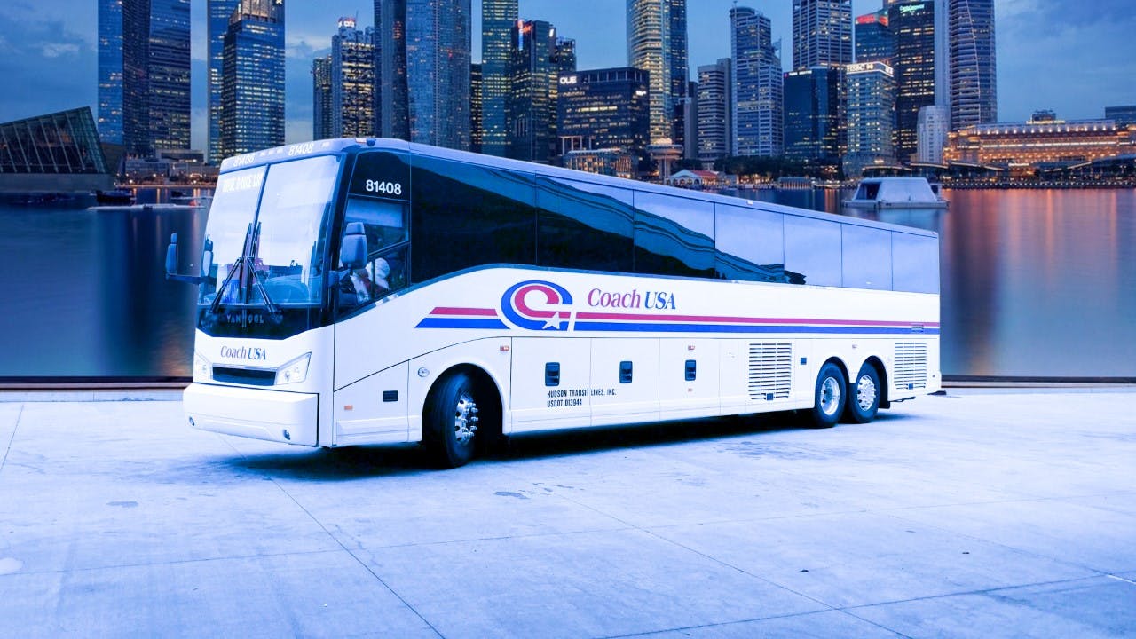 A white Coach USA bus parked on a concrete surface with a city skyline in the background.
