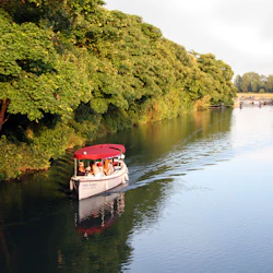 A small boat with a red canopy cruises on a river next to lush green trees. Passengers are seated inside.