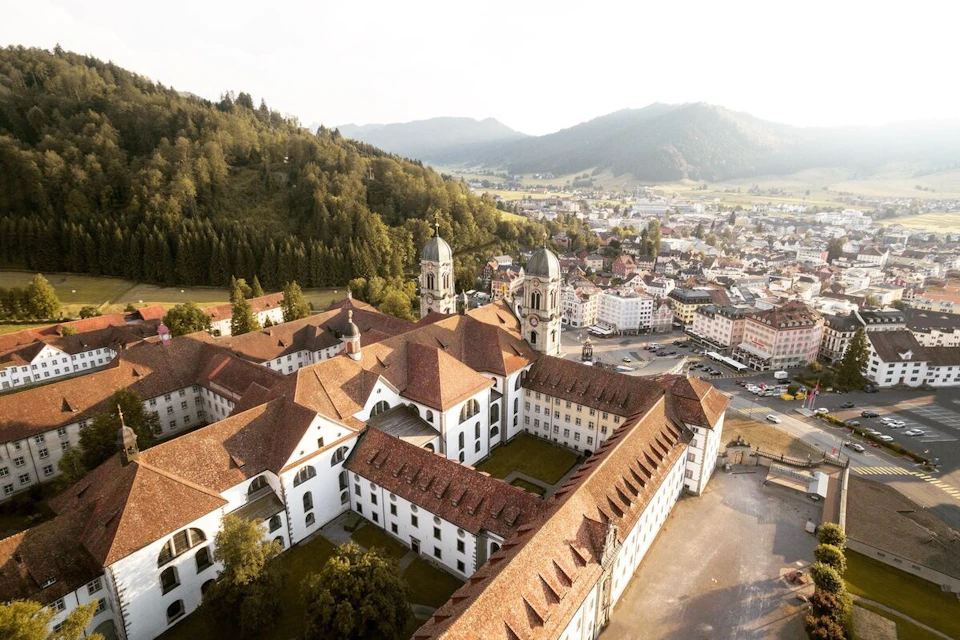 Aerial view of a historic building complex with red roofs, surrounded by a town, hills, and a forested landscape.