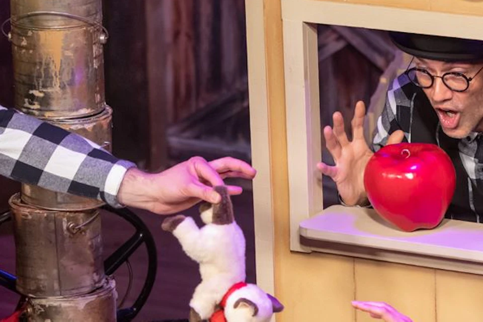 Three performers in a playful scene: one on a ladder, one in a window with an apple, and one reaching out with a red wig.