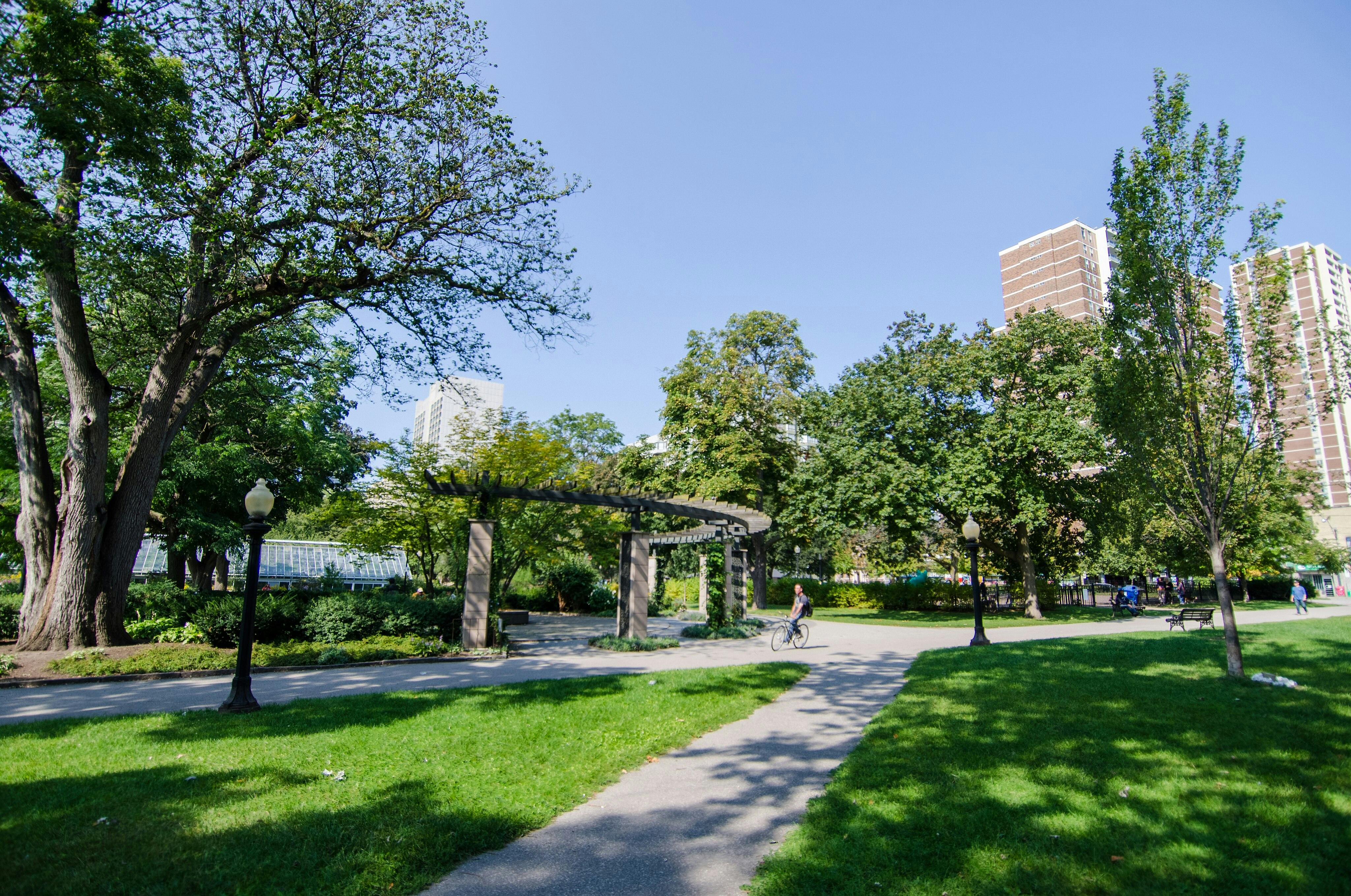 A sunny park with trees, green lawn, a pergola, path, lamp posts, a cyclist, and high-rise buildings in the background.