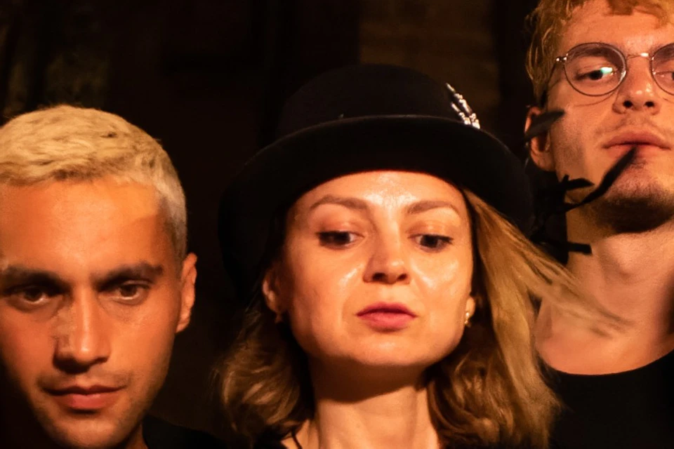Five people dressed in dark clothing, illuminated by a lantern, stand closely together at night with a Gothic building behind them.