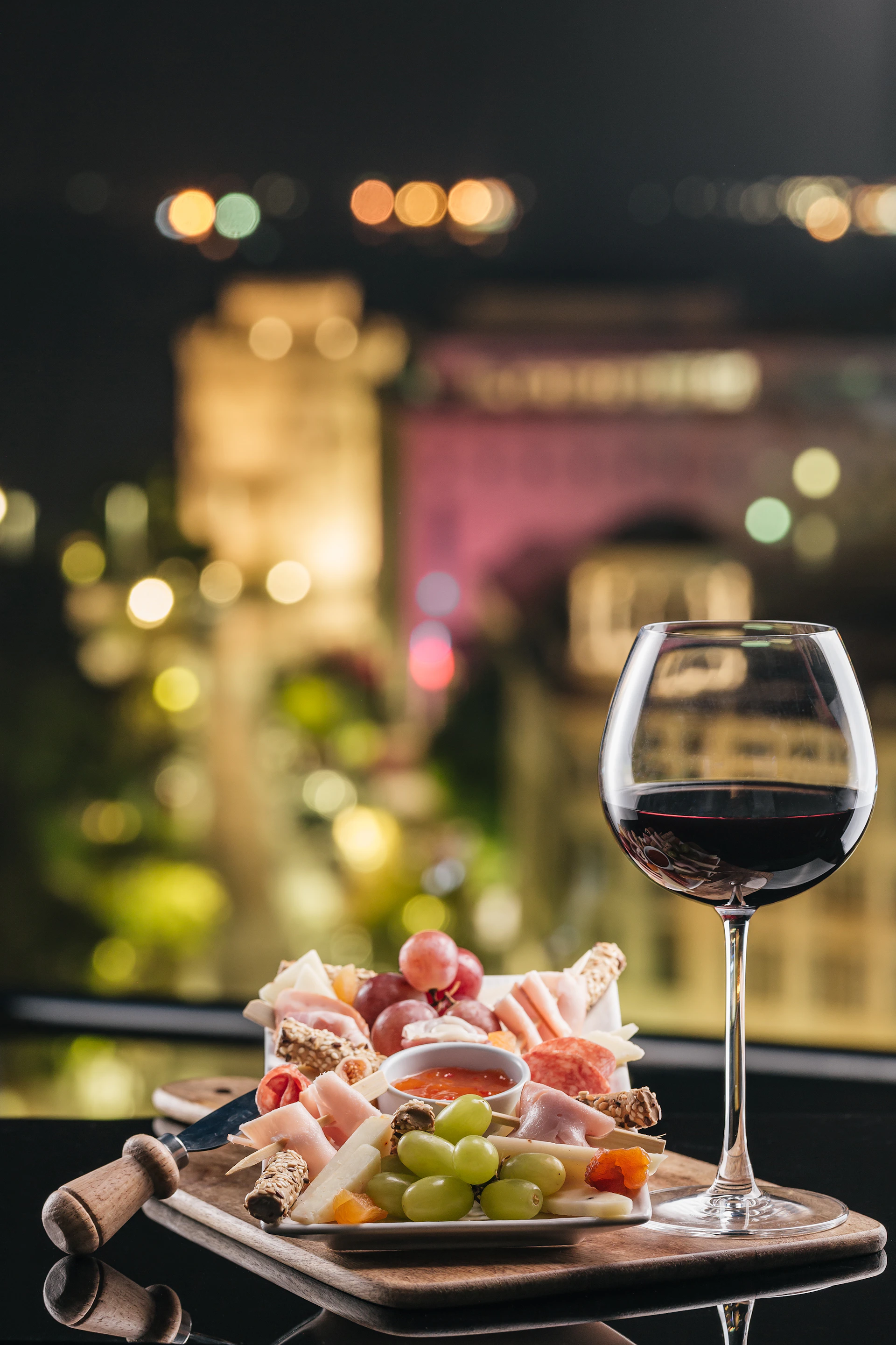 A glass of red wine and a charcuterie board with meats, cheeses, grapes, and crackers in front of a blurred cityscape at night.