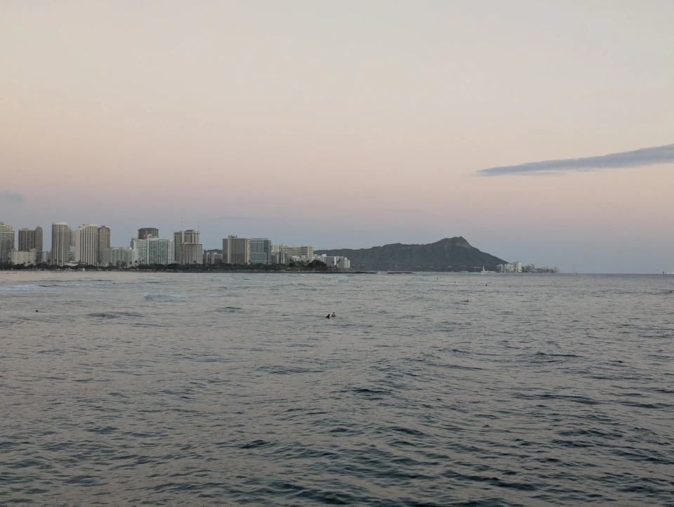 A view of a coastal city skyline with a mountain in the background, seen over a calm ocean under a pastel sky.
