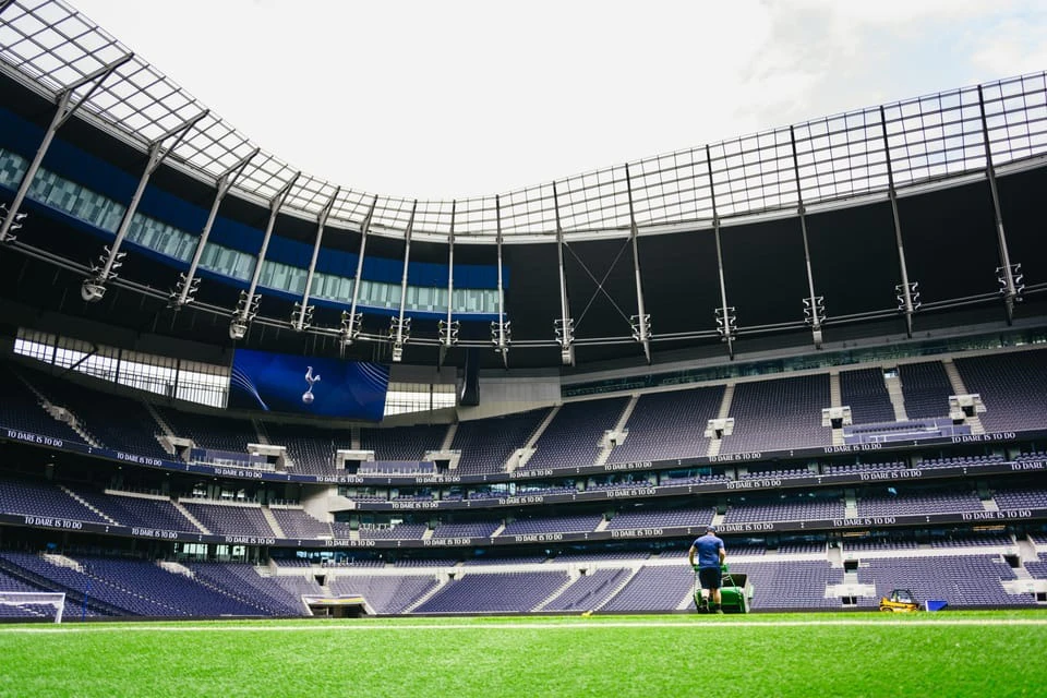 A person operates a lawn mower on a bright green field inside a large, empty stadium with tiered seating and a partial roof.