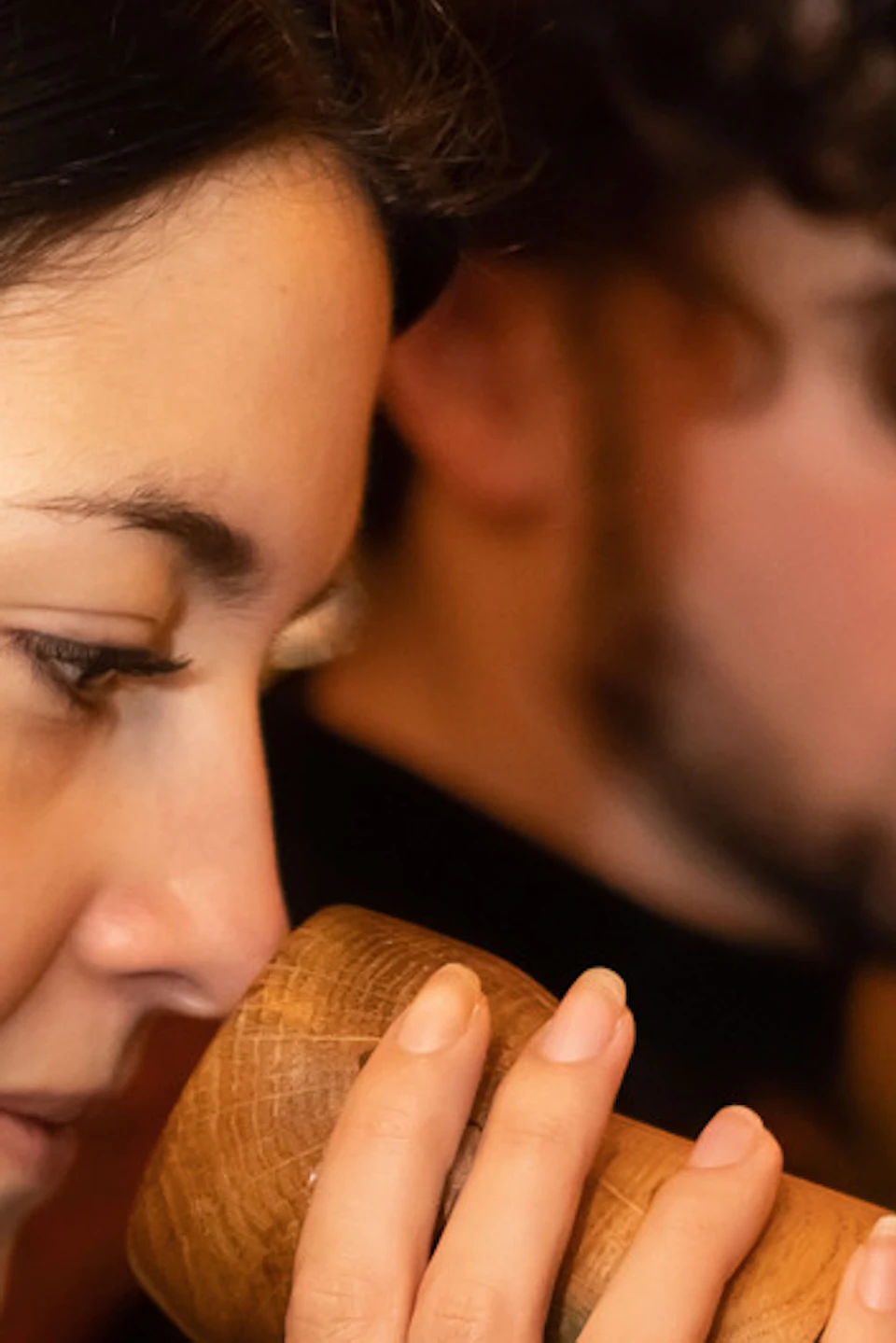 A woman smells a wooden container while a man stands in the blurred background in a warmly lit setting.