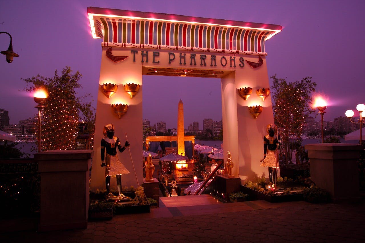 Vstup do zábavního parku nebo atrakce se staroegyptskou tématikou, světelnými nápisy, sochami a dekoracemi za soumraku.