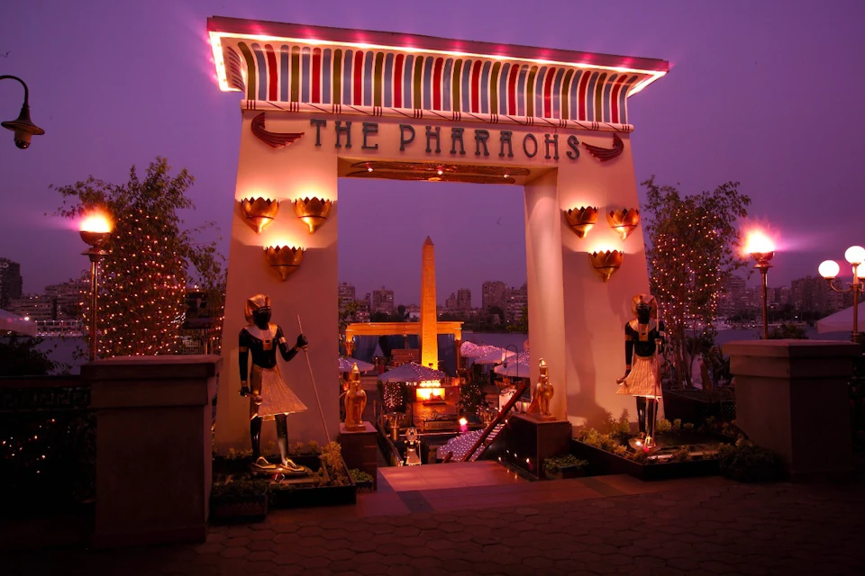 Entrance to a theme park or attraction with an ancient Egyptian theme, illuminated signs, statues, and decorations at dusk.