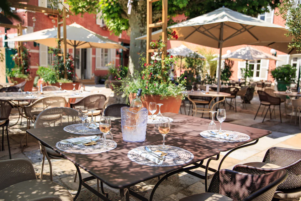 Outdoor restaurant patio with tables set with glassware, plates, and napkins under large umbrellas and surrounded by plants and trees.
