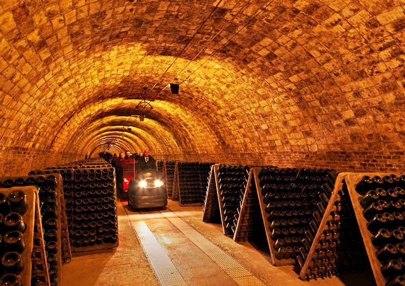 Des personnes circulant à bord d'un petit véhicule électrique traversent une cave à vin en briques voûtée, bordée de casiers à bouteilles.