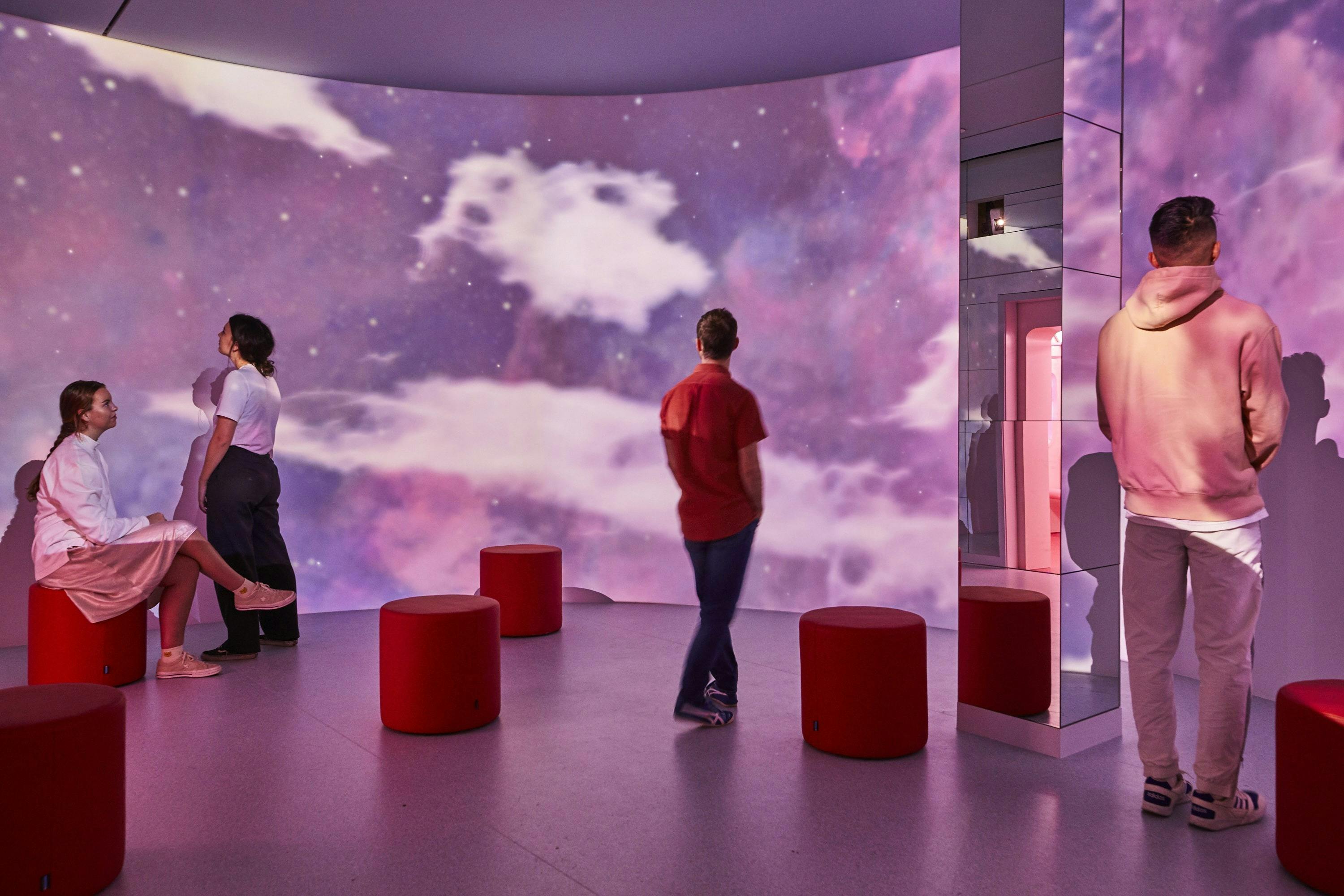 Tři lidé v místnosti s červenými stoličkami, kteří se dívají na velkou obrazovku zobrazující zataženou hvězdnou oblohu.