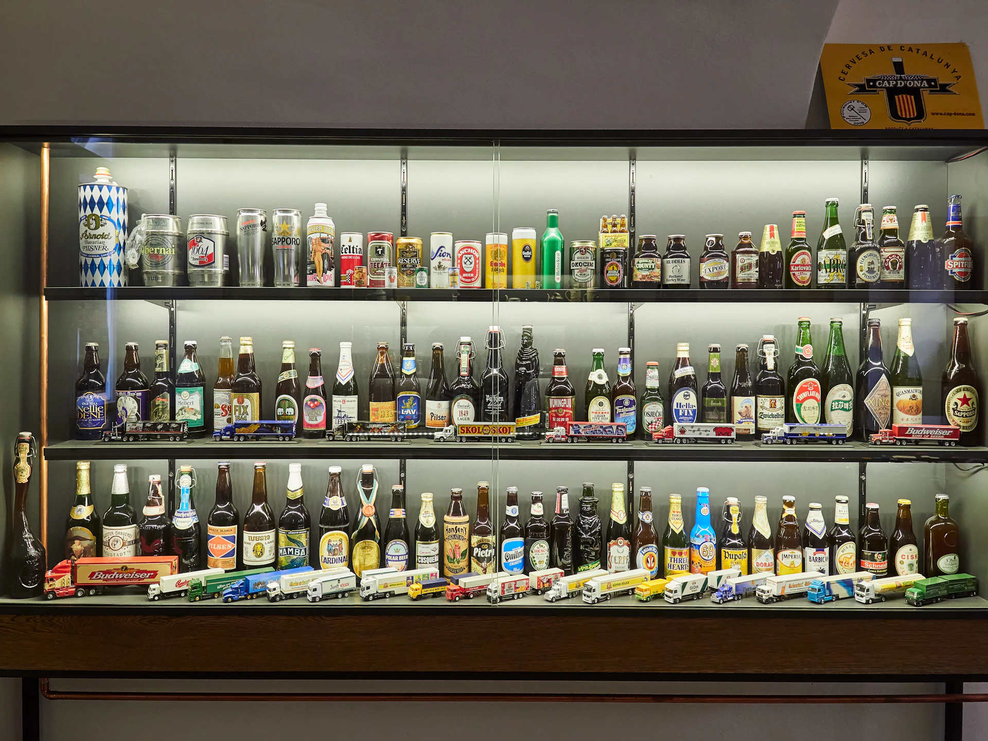 A display case with three shelves containing various bottles and cans of beer, along with miniature trucks at the bottom.