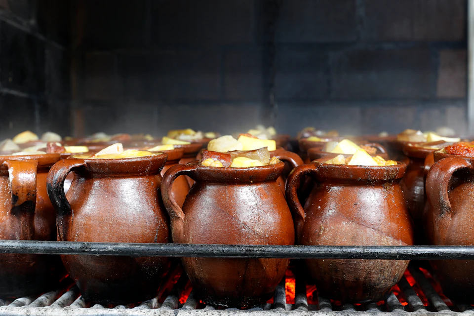 Several brown clay pots filled with chunks of vegetables are being cooked over a grate with red-hot coals visible below.