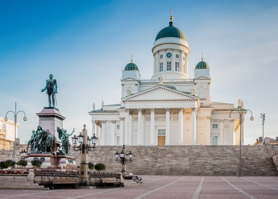 Large white cathedral with green domes, flanked by statues and ornate lampposts, against a blue sky.