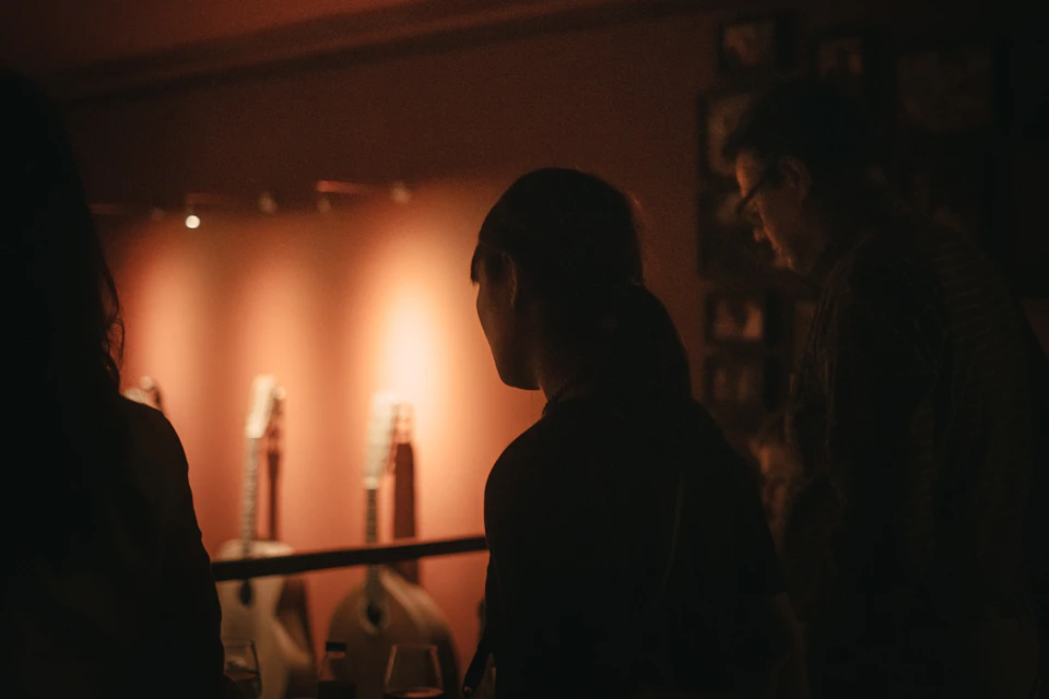 People viewed from behind in a dimly lit room with acoustic guitars displayed against a warmly lit wall in the background.