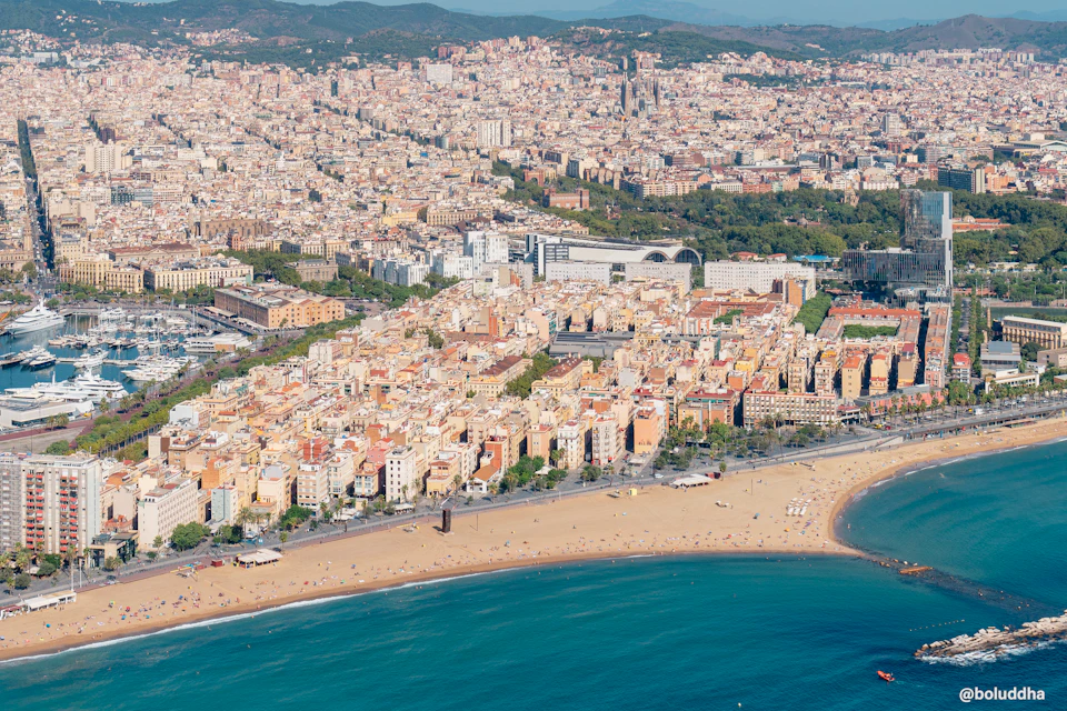 Aerial view of a coastal city with dense buildings, a wide sandy beach, and blue ocean water. Sparse people are visible on the beach.