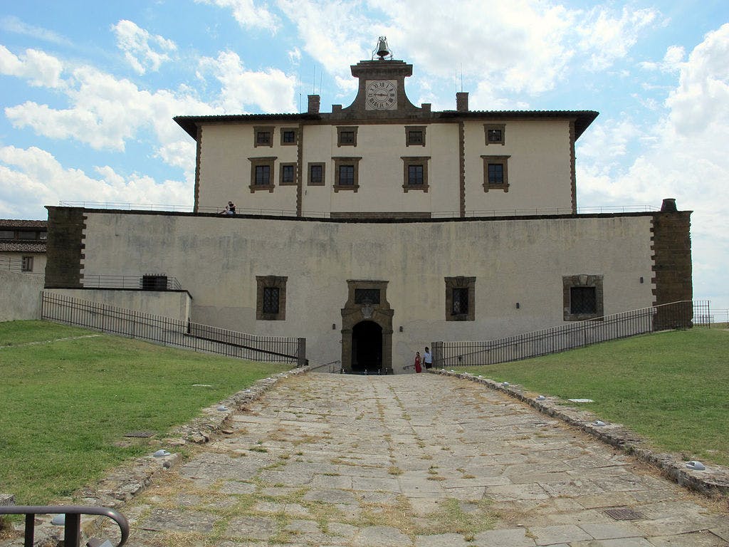 Old stone building with clock tower, surrounded by a high wall. Sloped stone pathway leading to entrance. Few people visible.