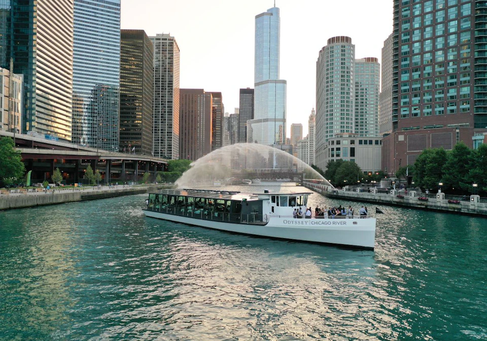 A boat named "Odyssey Chicago River" sailing on a river, surrounded by tall buildings and a water fountain arching over.