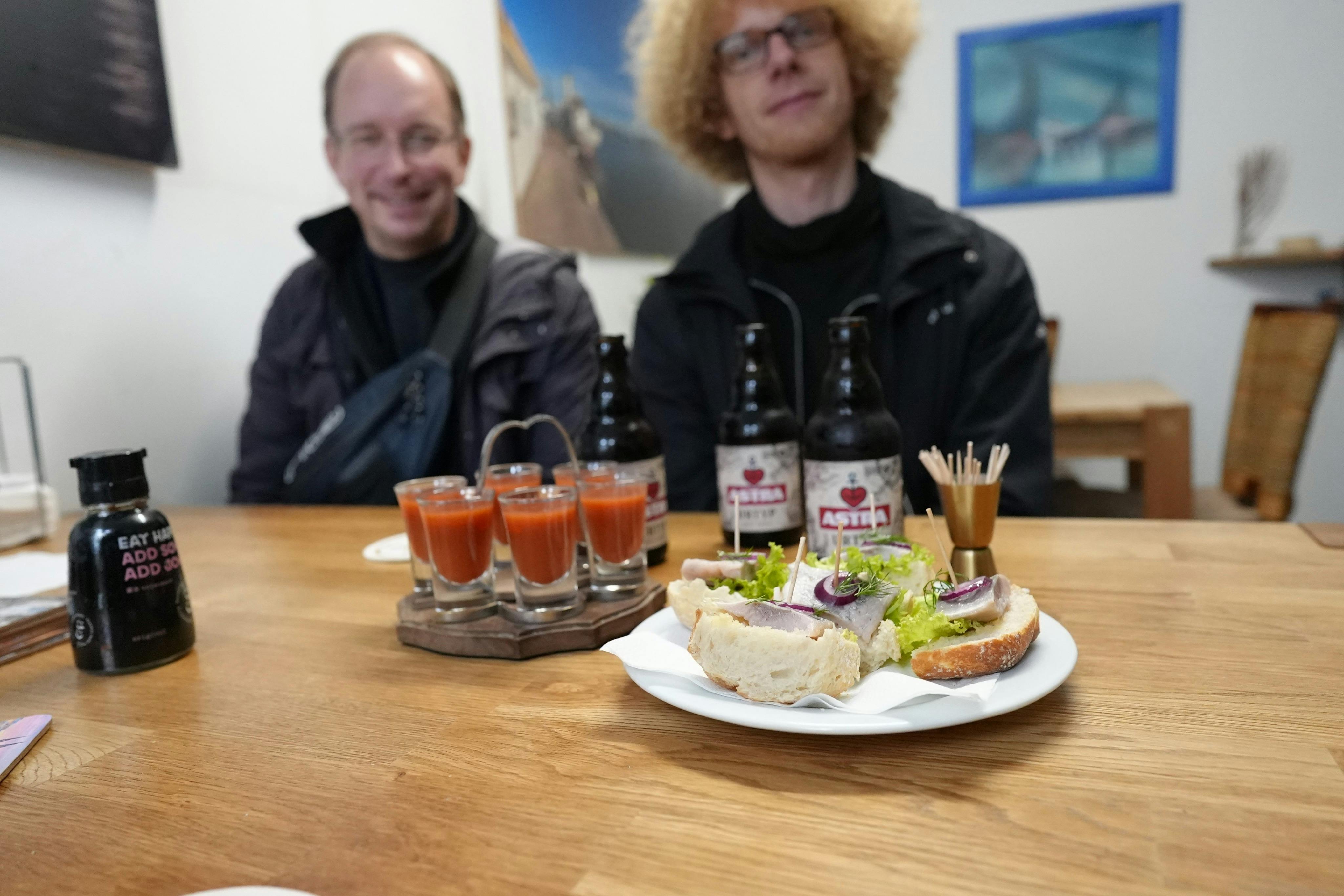 To personer sidder ved et bord med sandwich, tomatsaftshots og flasker øl. Kunst på væggene i baggrunden.