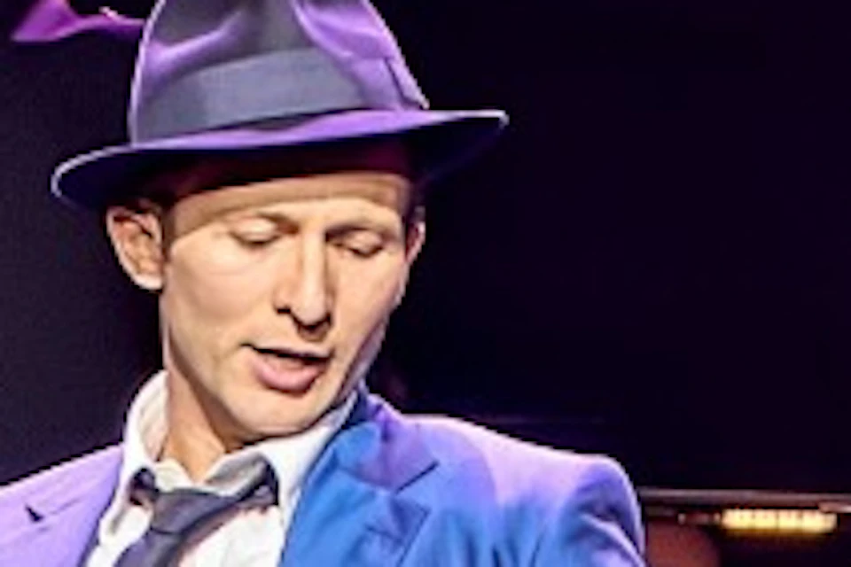 Performers in suits and hats dancing in unison on a stage under purple lighting, with a live band in the background.