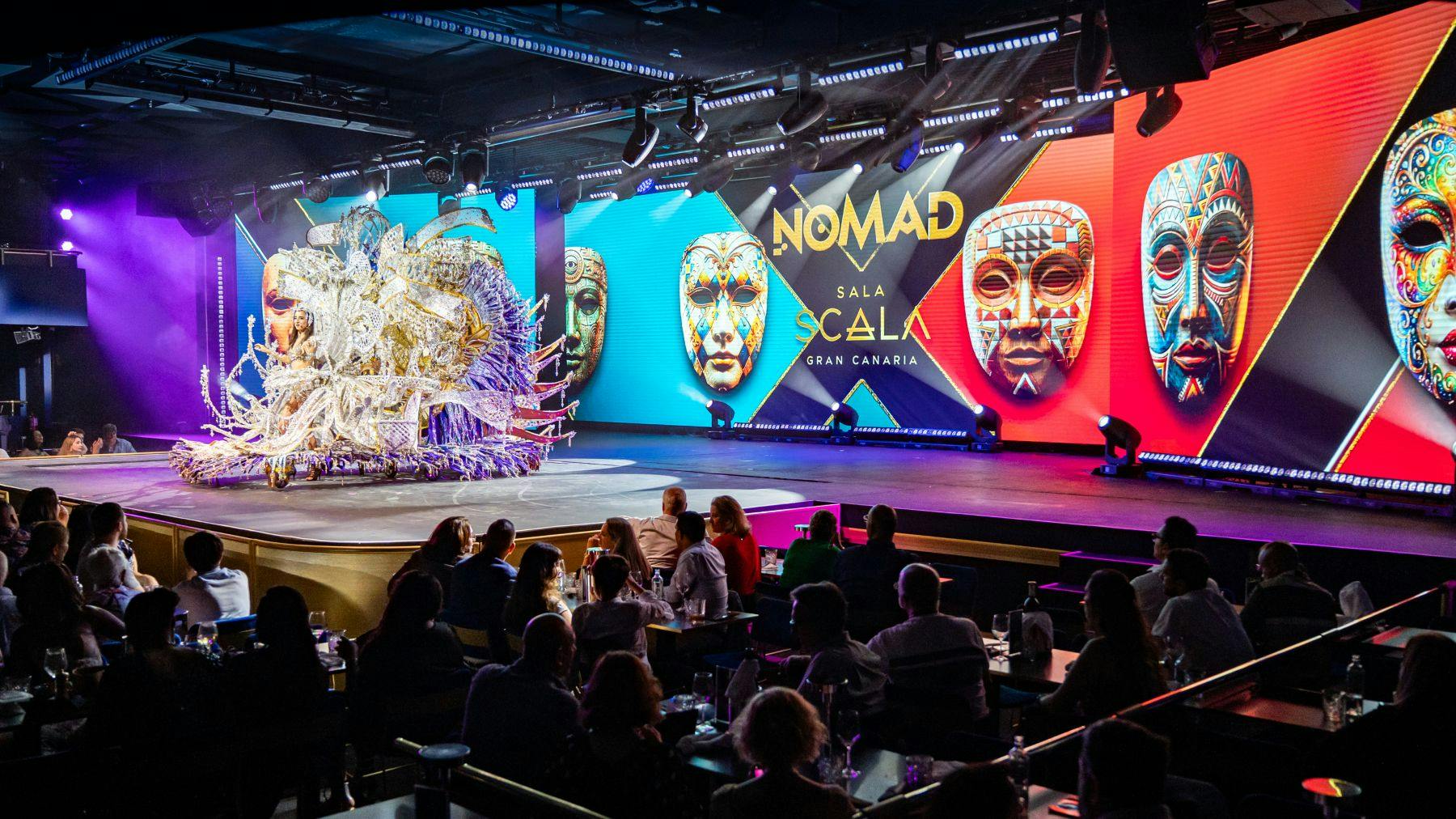 Um palco interior com máscaras coloridas exibidas em ecrãs onde se lê "Nomad Sala Scala Gran Canaria", com um público sentado.
