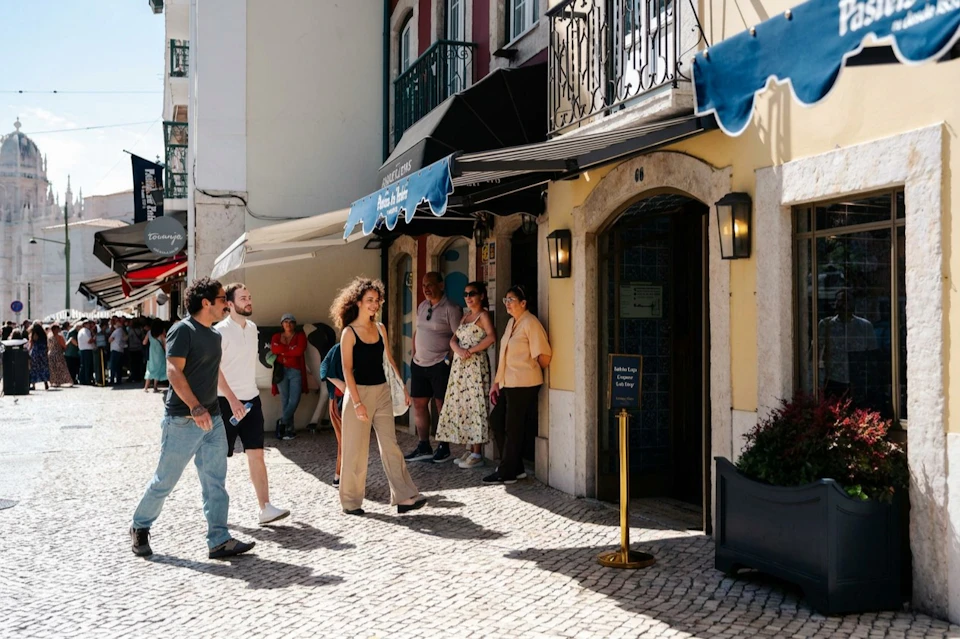 People walking and standing outside shops on a cobblestone street under awnings in a sunny urban setting.