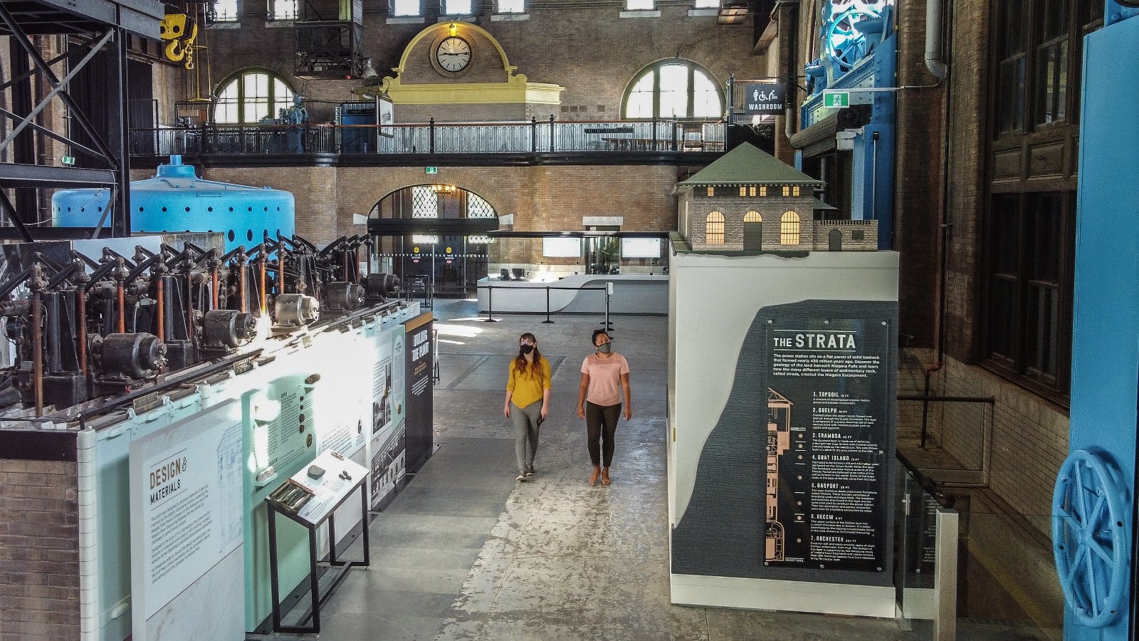 To personer, der går på et industrimuseum med forskellige udstillinger, herunder en stor plakat med titlen "LAGENE" og maskiner.