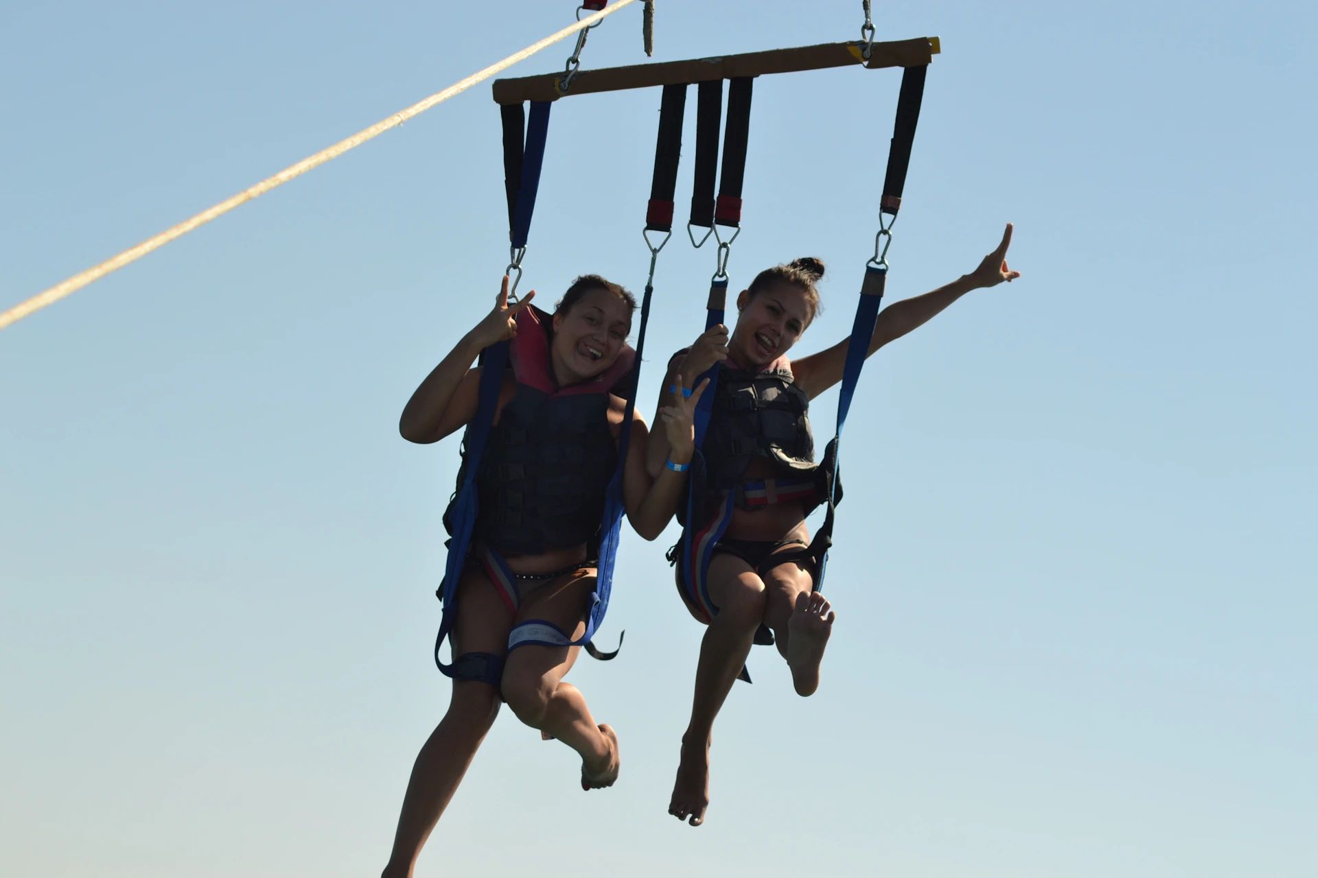 Two people smiling and wearing harnesses while parasailing against a clear blue sky. One person is pointing upwards.