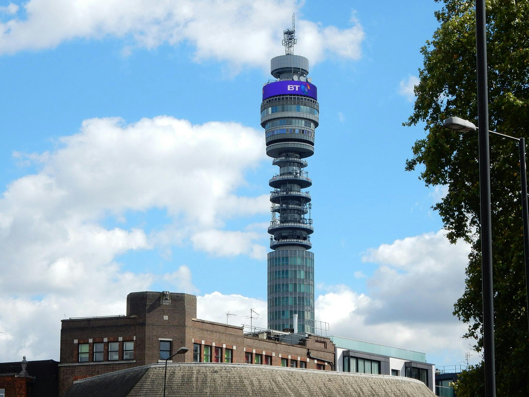 The BT Tower rises above buildings in a cityscape, under a blue sky with scattered clouds and next to a leafy tree.
