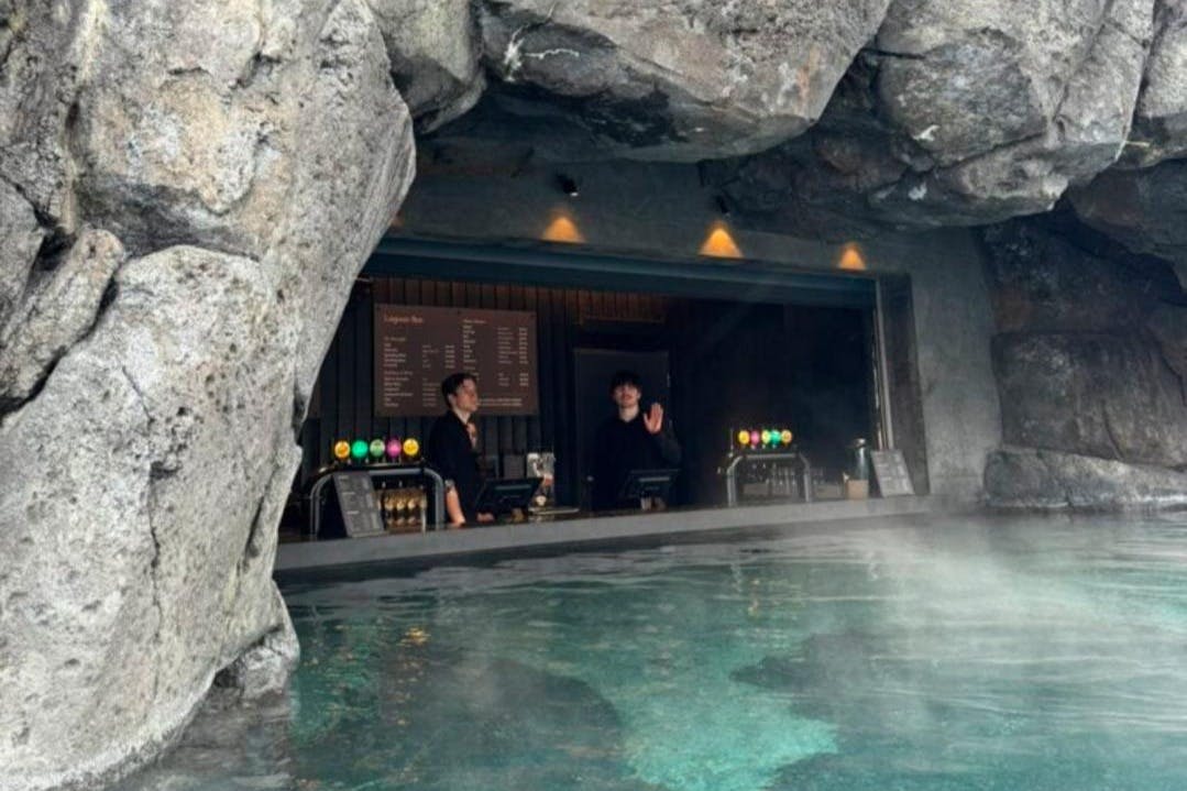 Two bartenders stand behind a bar set inside a rocky cave, with a pool in the foreground and a menu on the wall behind them.