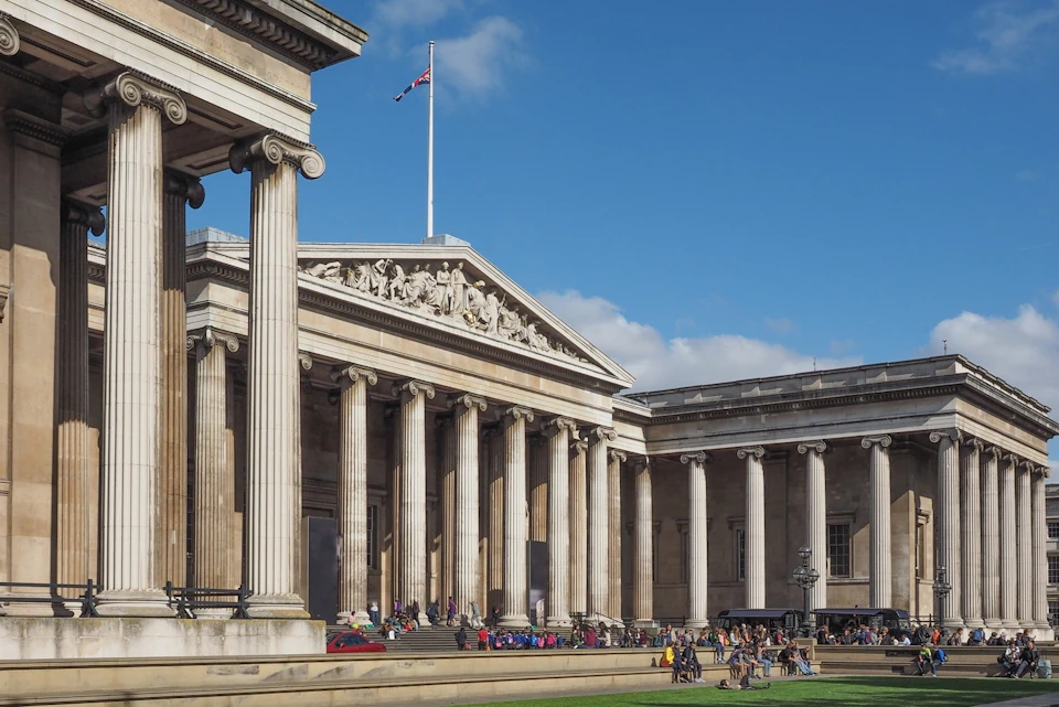 A neoclassical building with columns and a triangular pediment, featuring people gathered in the foreground under a blue sky.