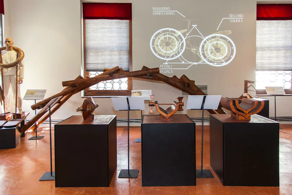 Exhibit with wooden sculptures on pedestals, annotated bicycle diagrams projected on the wall, and a window with red and grey curtains.