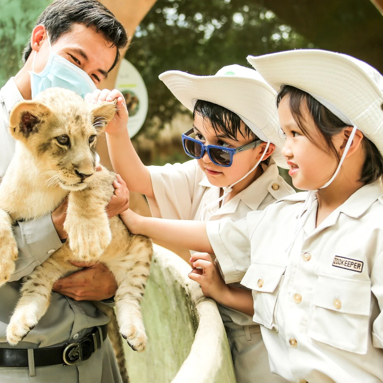 A zookeeper holding a lion cub while two children in uniforms and hats interact with the cub at a zoo.