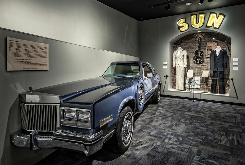 A blue classic car is displayed in a museum near a mannequin in a white suit and a guitar. A "SUN" neon sign is partially visible.