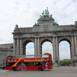 Röd dubbeldäckad sightseeingbuss framför ett stort valvmonument med statyer, under en molnig himmel i Bryssel.