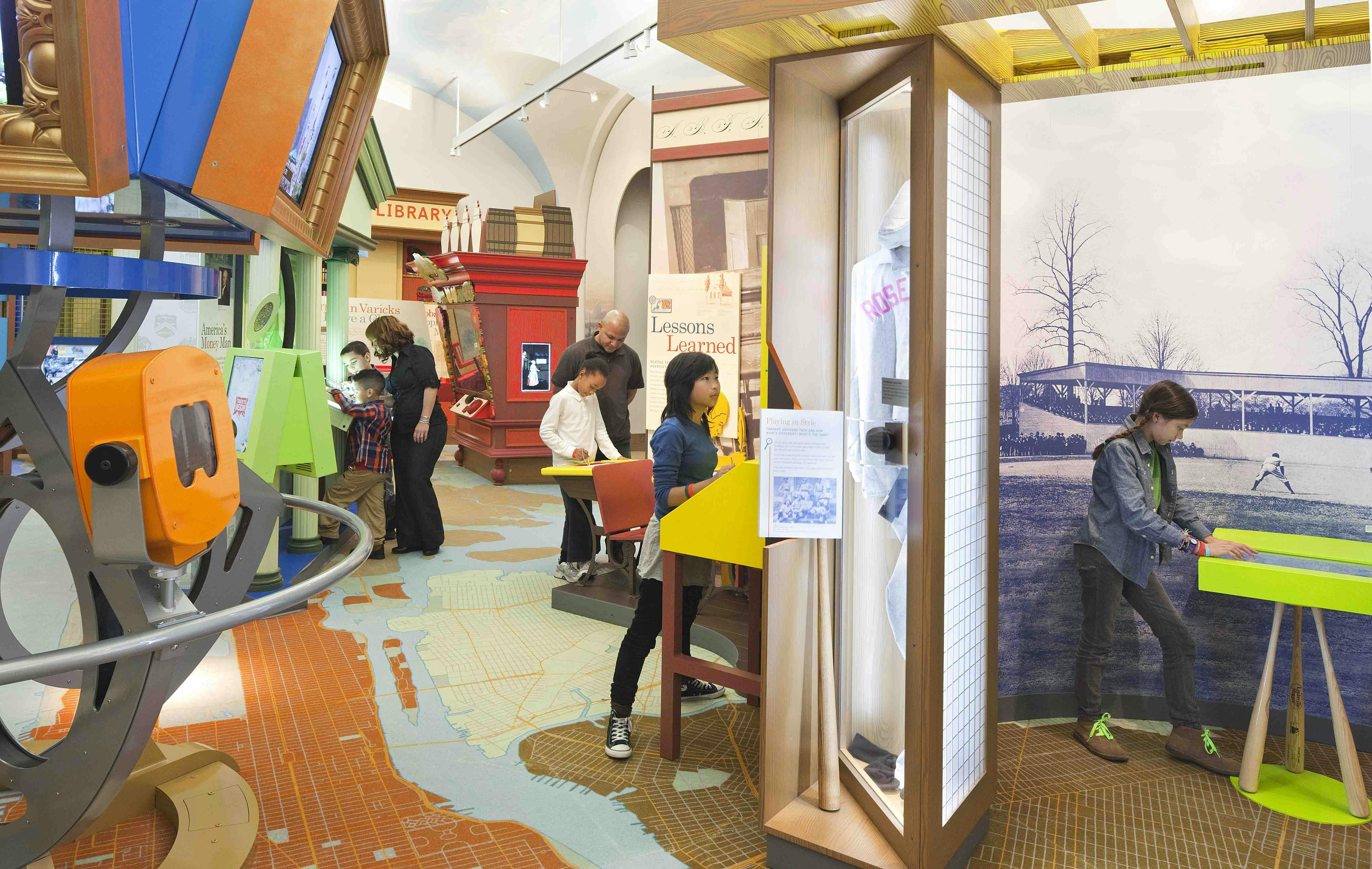 Παιδιά και ενήλικες εξερευνούν διαδραστικά εκθέματα σε ένα πολύχρωμο, εκπαιδευτικό μουσειακό περιβάλλον, με χάρτες, οθόνες και ιστορικά αντικείμενα.