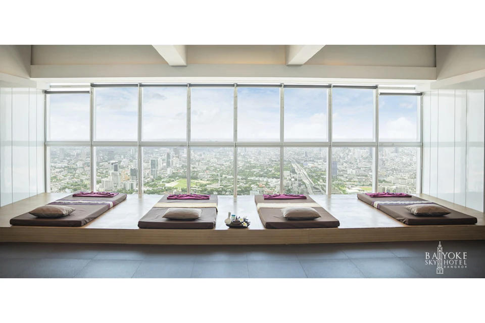 A spa room with elevated platforms and cushions, overlooking a cityscape through large floor-to-ceiling windows.